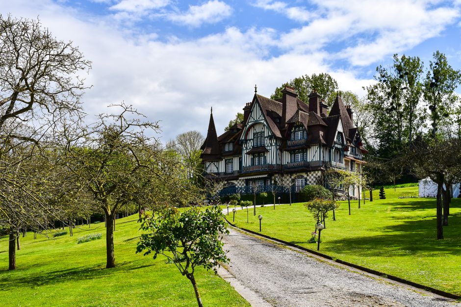 A large, historic Victorian-style house with intricate Tudor Revival architectural details, situated amidst a well-maintained garden. In the foreground, a gravel pathway curves gently through a lush, 